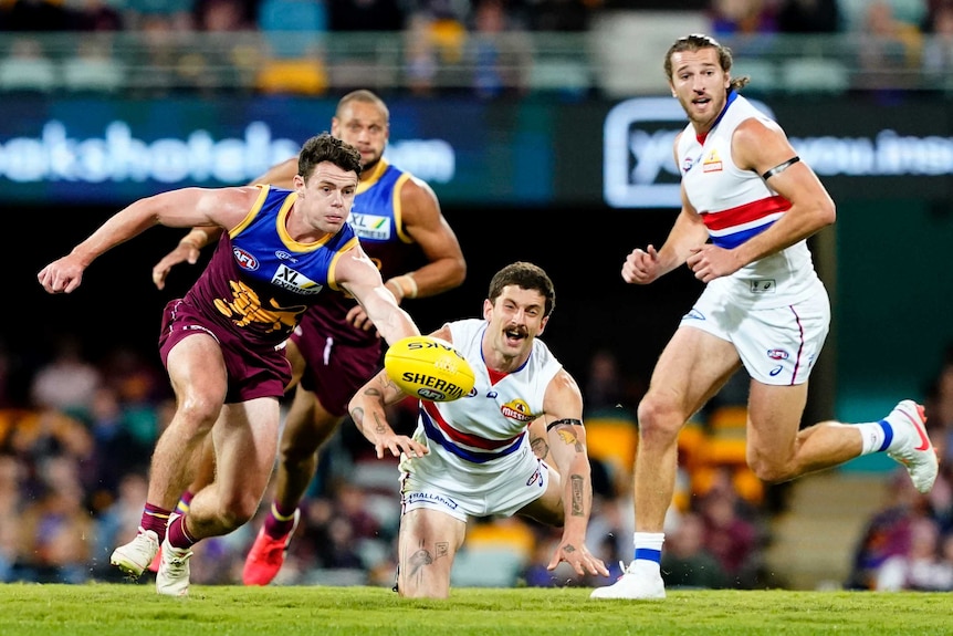 A Brisbane Lions AFL players runs for the ball as two Western Bulldogs opponents look on.