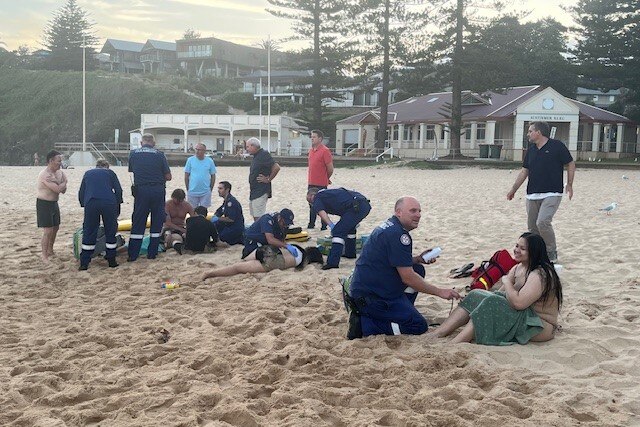 A paramedic on a beach attends to a seated person with a cluster of people behind