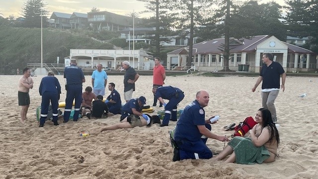 A paramedic on a beach attends to a seated person with a cluster of people behind