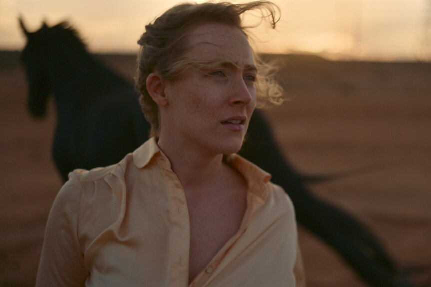 A blonde white woman in her 30s wearing a pale yellow shirt stands outside in a desert, with wind blowing.