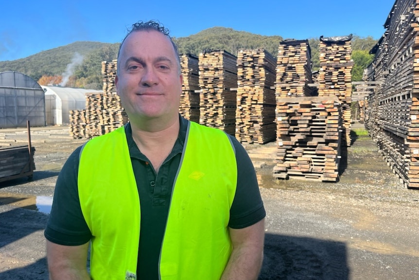 A middle-aged man in a high-vis vest standing in front of piles of wood.