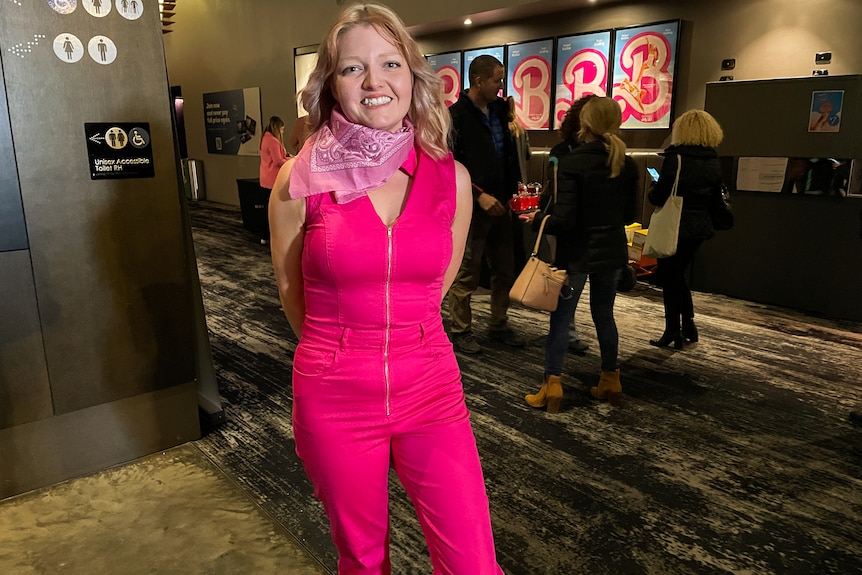 Jenny smiles in a cinema foyer in front of Barbie posters, wearing a hot pink cowgirl set.