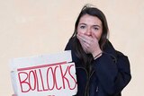 A woman holds a hand over her mouth while hiding a sign that reads Bollocks to Brexit