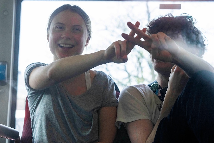 A young woman sits next to a young man on a bus making the peace sign with her fingers and smiling