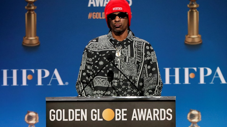 Snoop Dogg wearing a red beanie and black and white shirt and sunglasses at a lectern