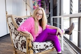 A white woman with blonde hair in her late 40s wears a bright pink shirt and purple leggings and sits in a cow hide chair.