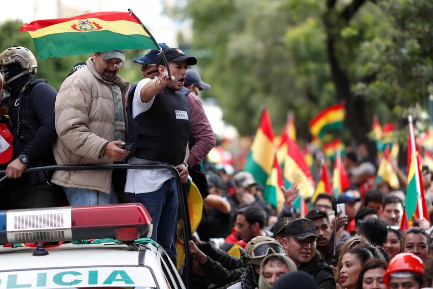Luis Fernando Camacho stands on the back of a police vehicle waving Bolivian flag.