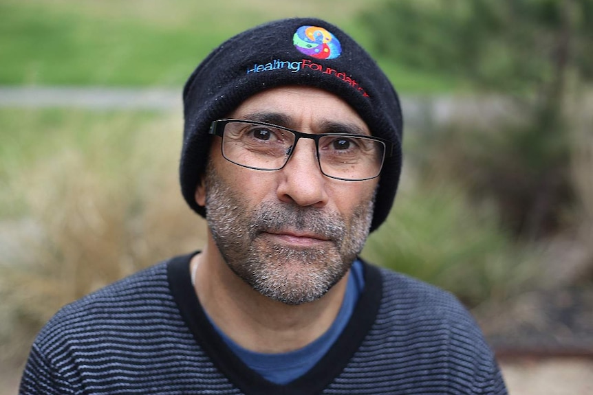 Ian Hamm, wearing a 'Healing Foundation' black beanie, looks contemplative as he sits in a park.