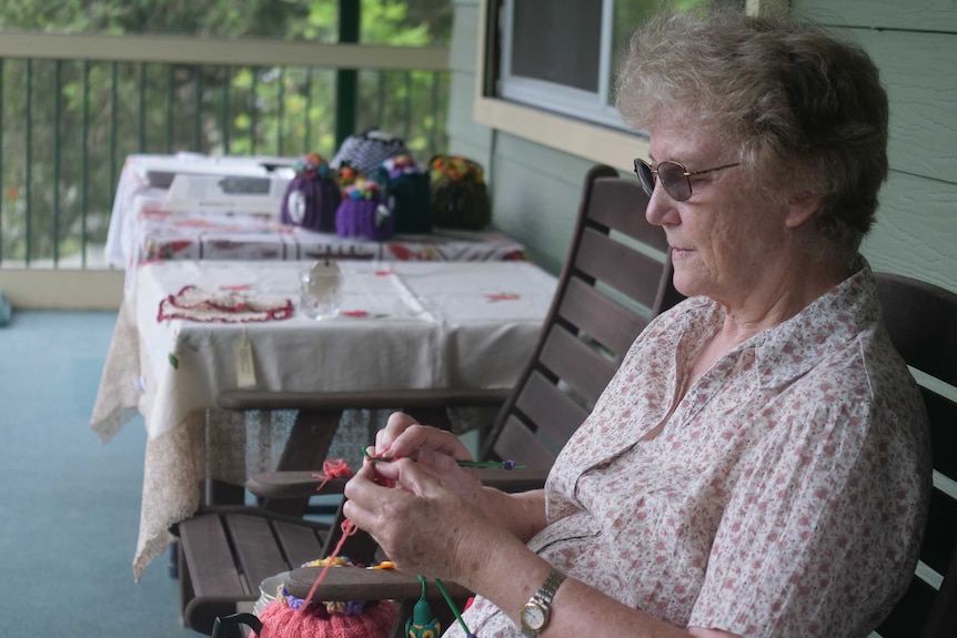 An older lady sitting on a chair knitting on a deck.