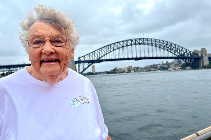 an elderly woman wearing glasses smiling with the harbour bridge behind her