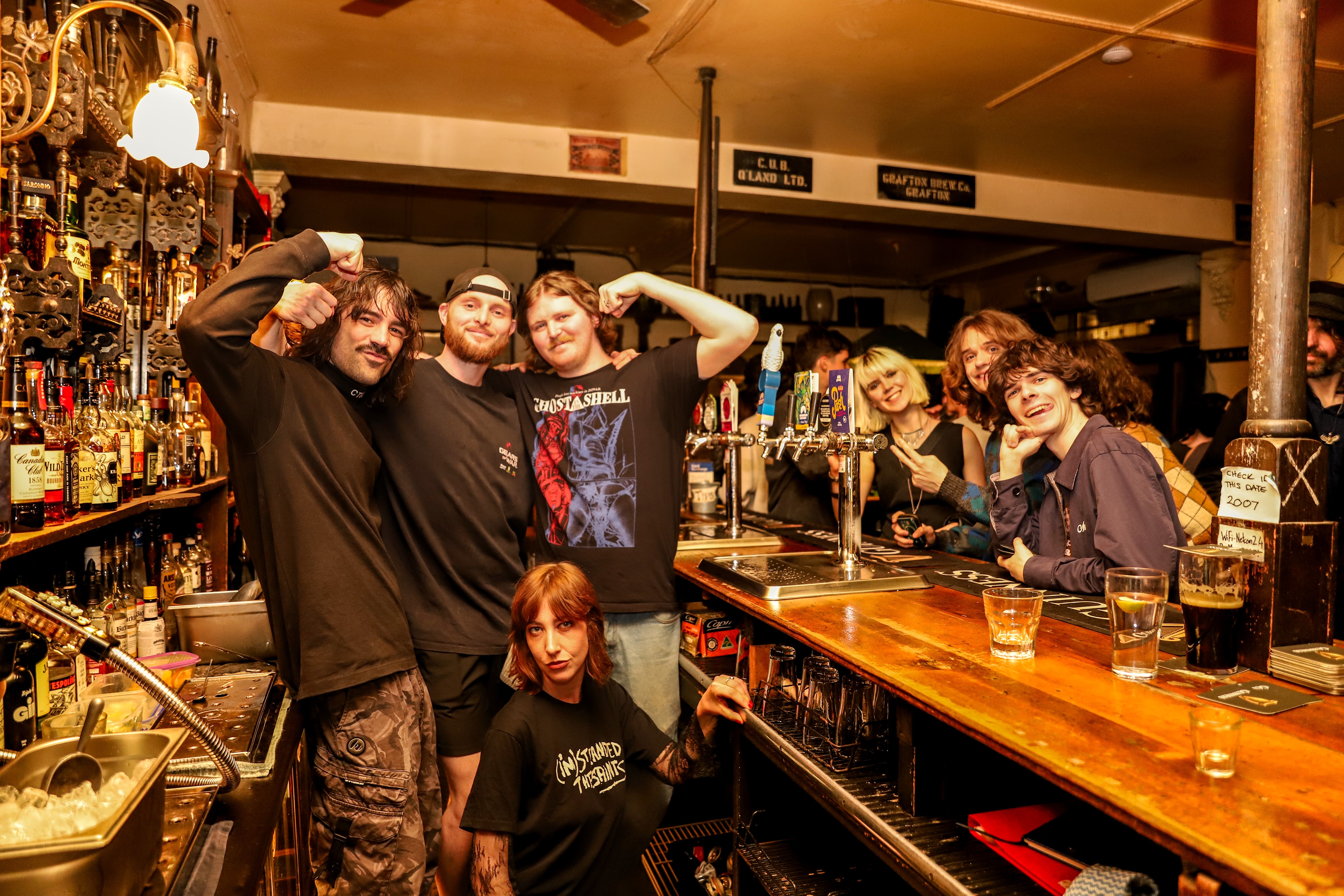 Quatro funcionários do bar sorriem e posam atrás do balcão de um bar movimentado no centro da cidade sob iluminação amarela