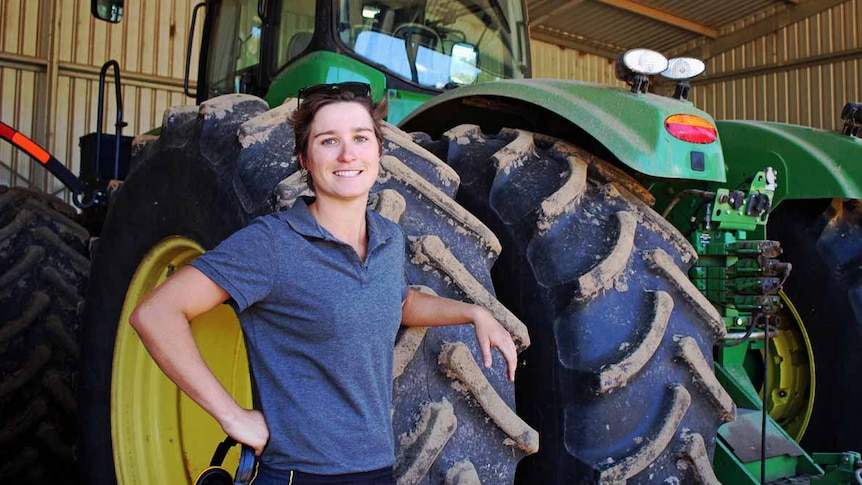 Woman smiles in a farm shed as she leans against the tires of a green tractor.