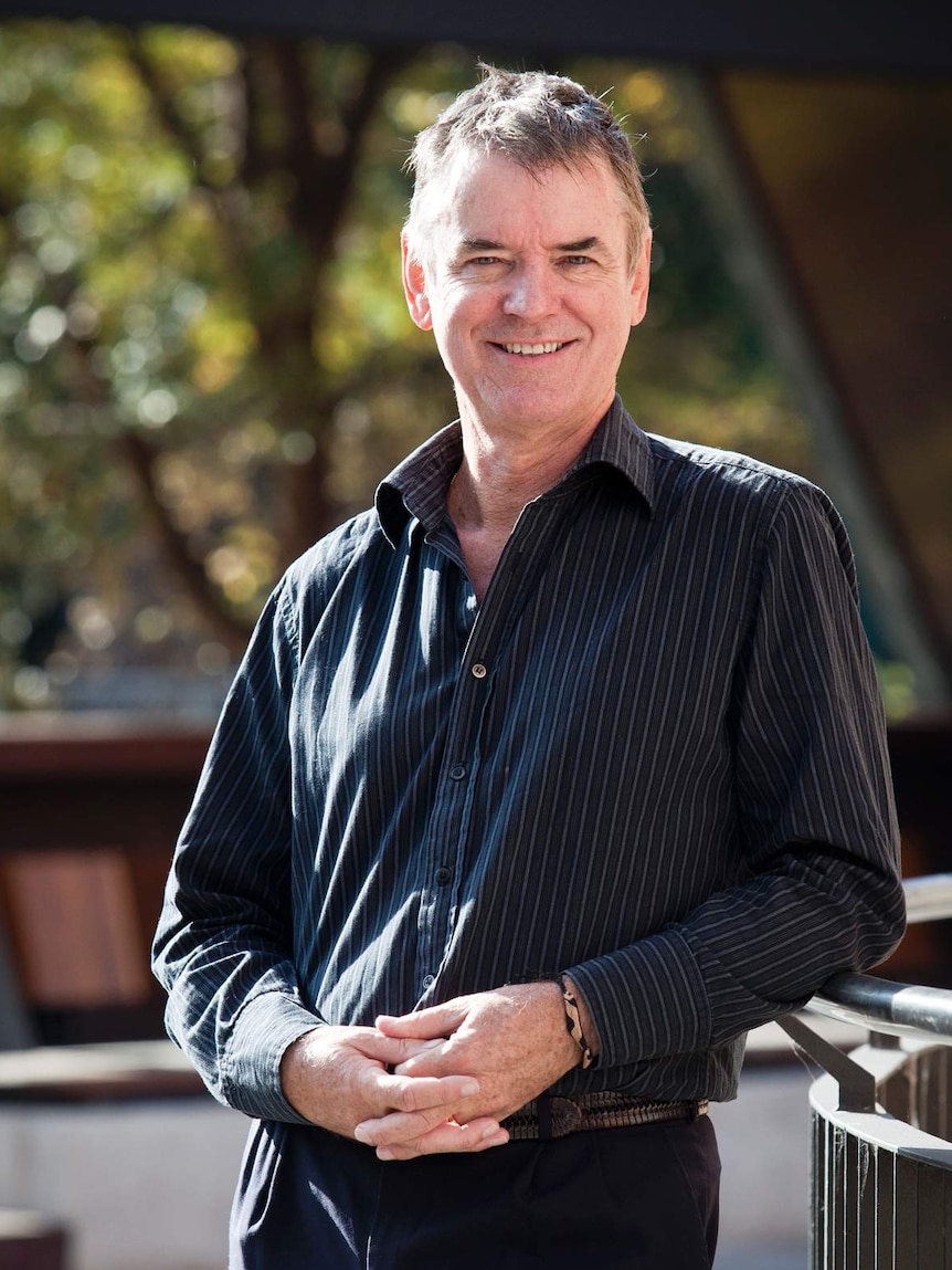 A man in a blue shirt and pants leans against a railing and smiles.