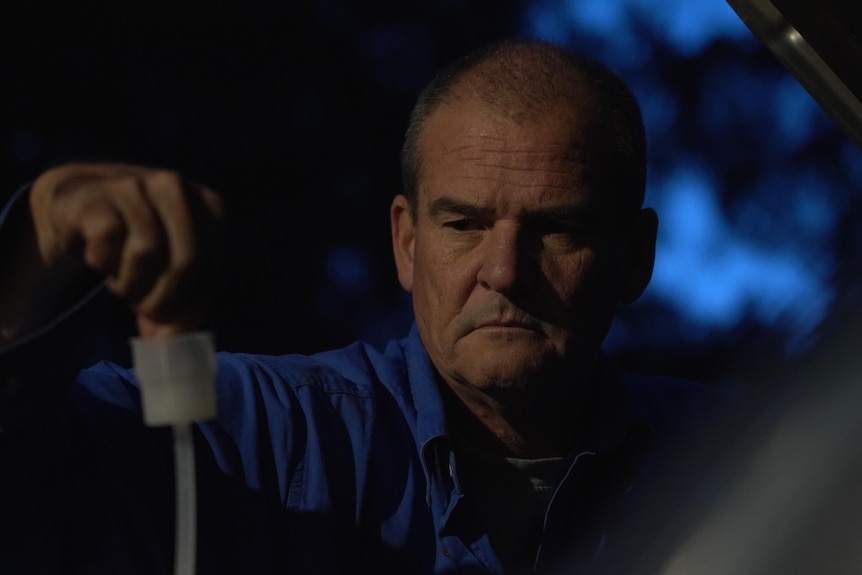 A man in a long-sleeve blue shirt looks downh slightly sitting in a darkened room