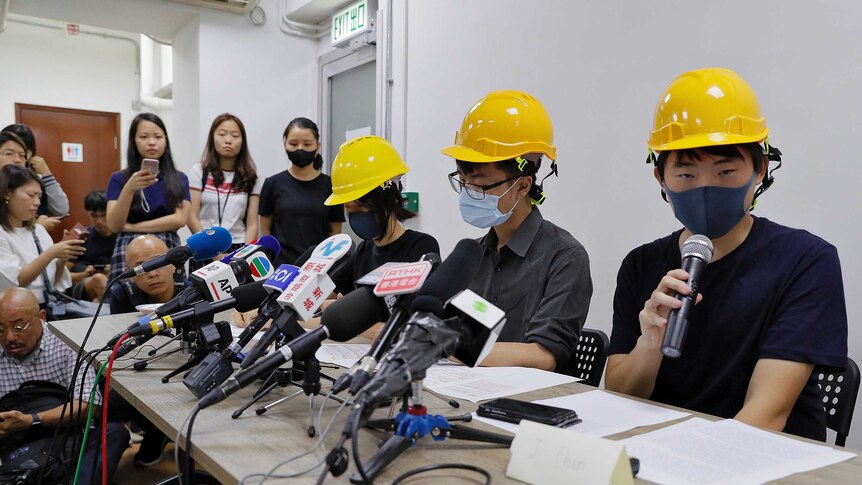 Three protesters wearing yellow hardhats and wearing face masks speak at a press conference in front of media microphones