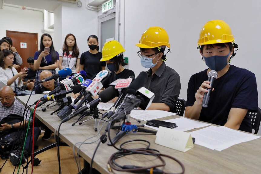 Three protesters wearing yellow hardhats and wearing face masks speak at a press conference in front of media microphones