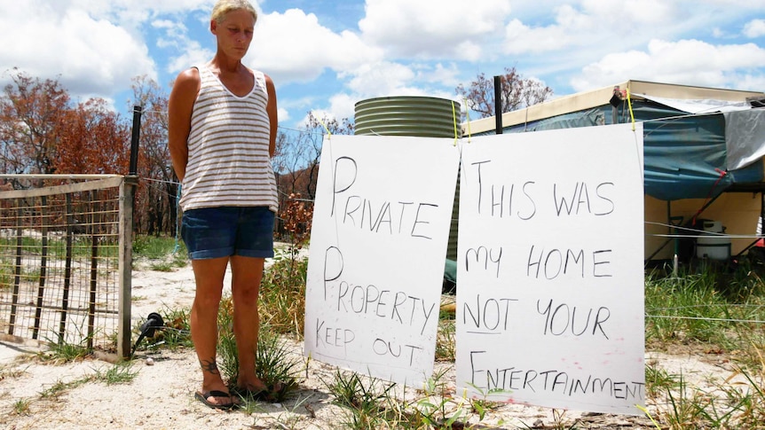 A middle aged woman stands solemnly next to a sign which says private property keep out, this was my home not your entertainment