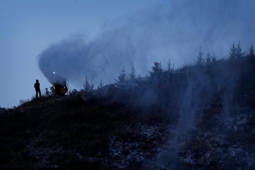 A person works at a snow making machine on a hill.