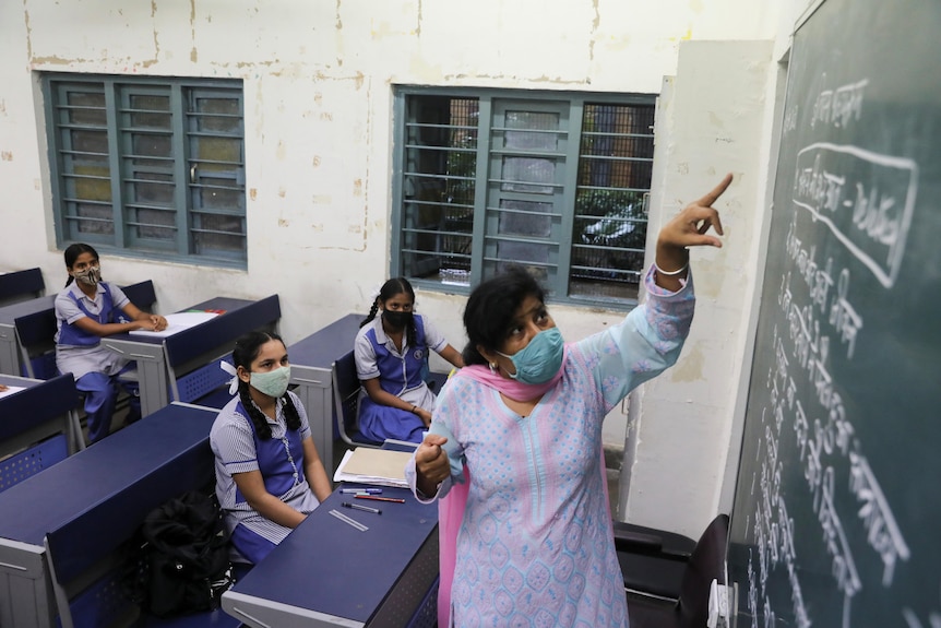 A teacher points at a blackboard while a few students in a mostly empty classroom look on. All wear masks. 