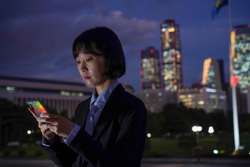 A woman with cropped black hair looks down at her phone with the city of Seoul lit up behind her.