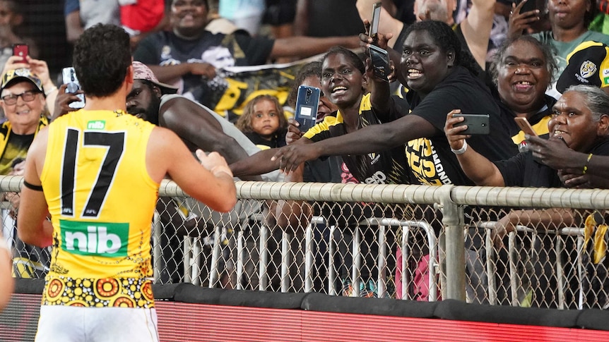 An AFL footballer gestures at the crowd as Indigenous fans smile and take pictures.