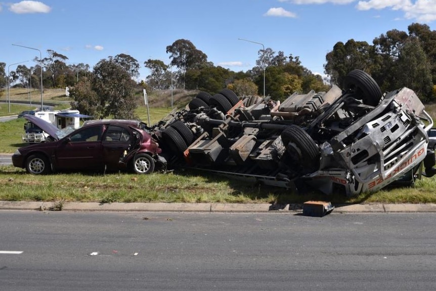 A crushed car and an upturned truck on the grass by the side of a road.