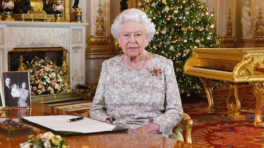 Queen Elizabeth II sits at a desk in  a grand drawing room with a Christmas tree behind her