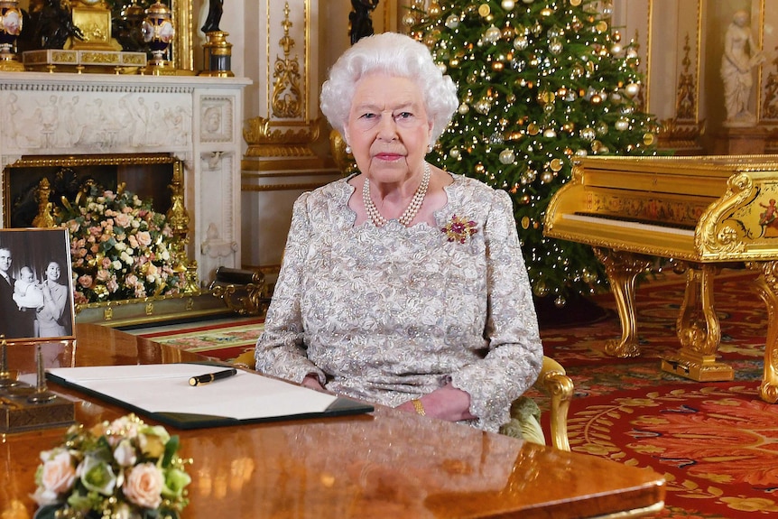 Queen Elizabeth II sits at a desk in  a grand drawing room with a Christmas tree behind her