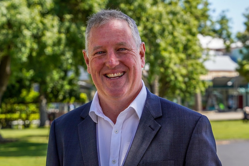 A man in a light collared shirt and navy suit jacket smiles in front a backdrop of green leafy trees.