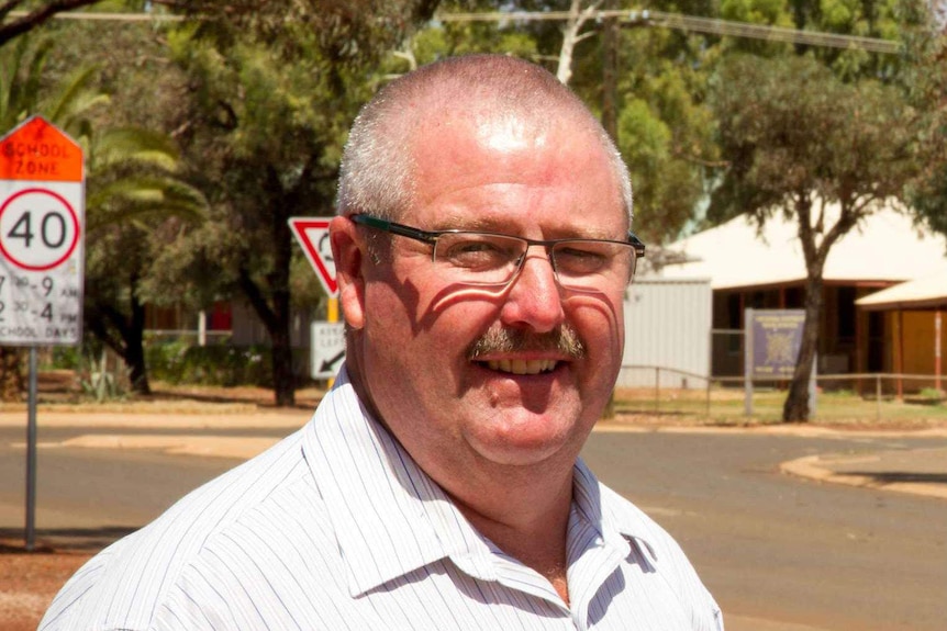 A man with grey hair, glasses and a moustache, wearing a business shirt, stands smiling and squinting in the sun, near a street.