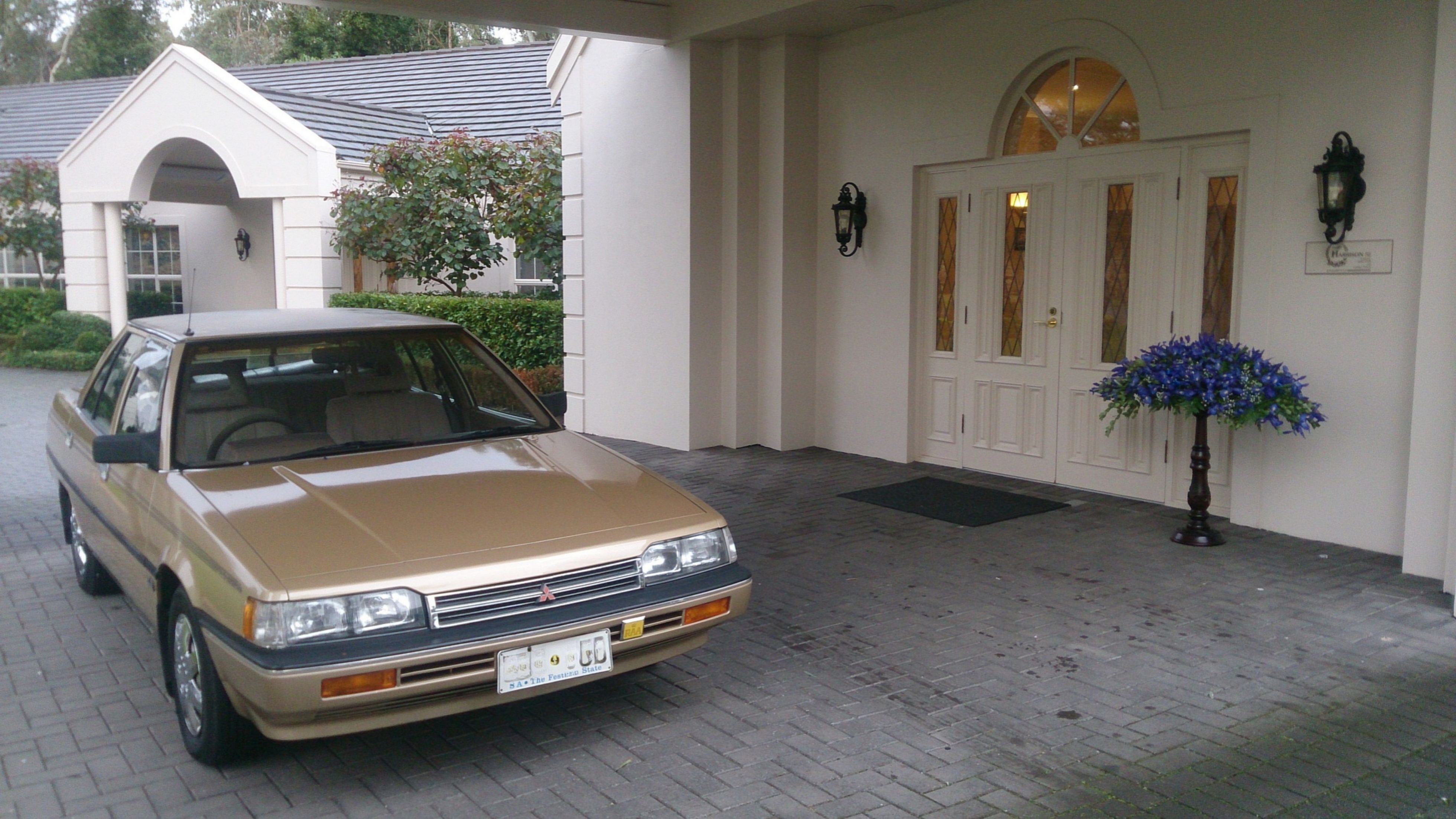 A gold-coloured car parked in the driveway of a white mansion.
