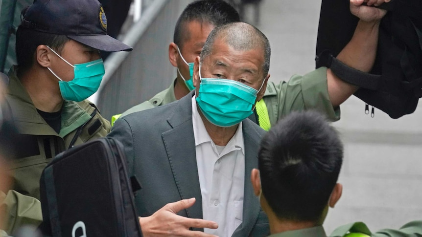 A man in a suit is surrounded by police wearing khaki uniforms, with all people pictured wearing disposable face masks.