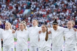 A women's soccer team wearing white sings with a crowd behind them