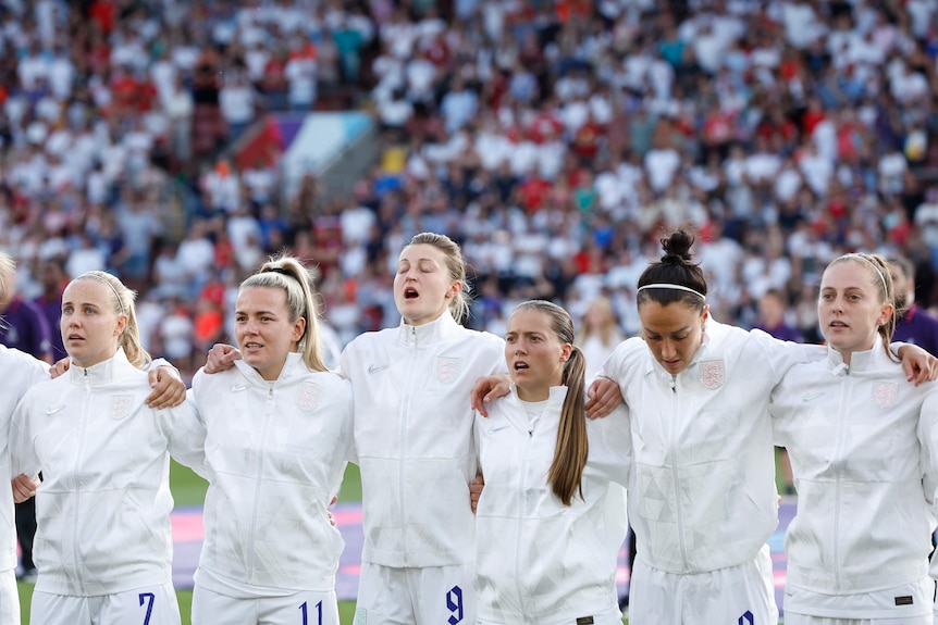 A women's soccer team wearing white sings with a crowd behind them