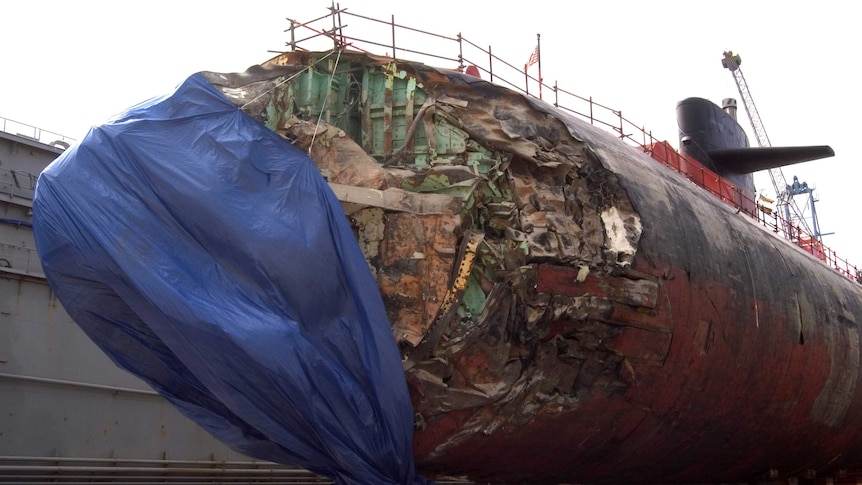 A US Navy submarine with a heavily damaged nose sits in dry dock.