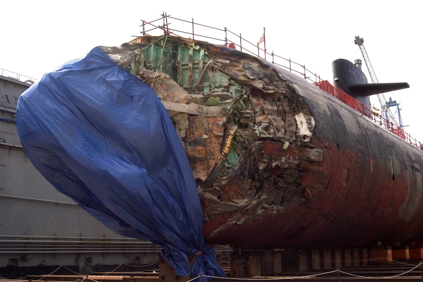 A US Navy submarine with a heavily damaged nose sits in dry dock.