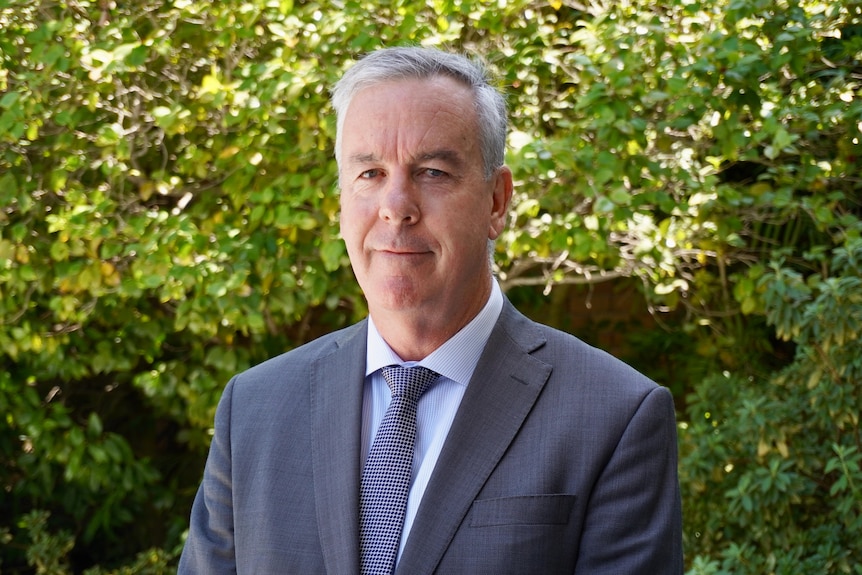 A man in a suit and tie stands with a slight smile. 