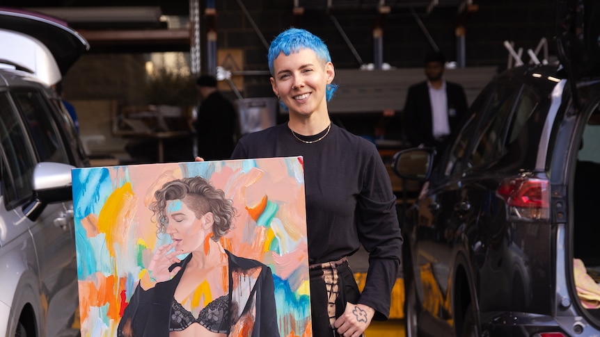 A white androgynous woman with bright blue hair stands in a carpark holding a colourful portrait of Virginia Gay.