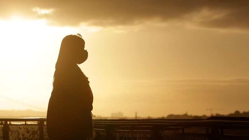 A woman wearing a face mask is silhouetted against the setting sun.
