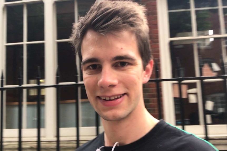 A young man stands on the footpath and looks into the camera.