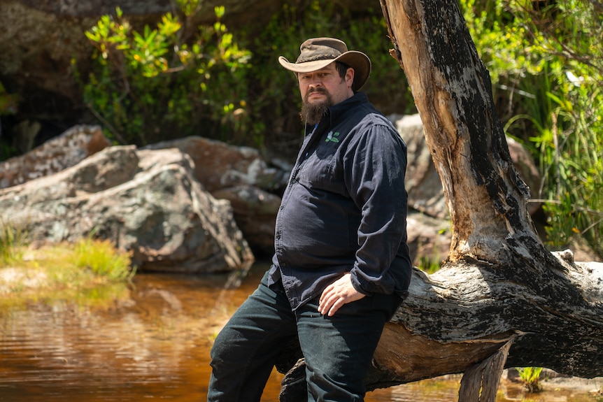 A man with a dark beard and shirt in an Akubra stares at the camera. he is sitting on a tree branch on a flowing creek bed.