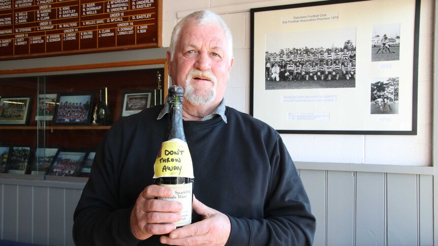 A man holds a dusty bottle of champagne in front of old football photos.