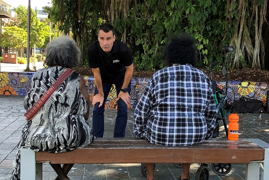 Young man speaks to two Indigenous Australian women sitting on wooden bench in Cairns with large tree in background.