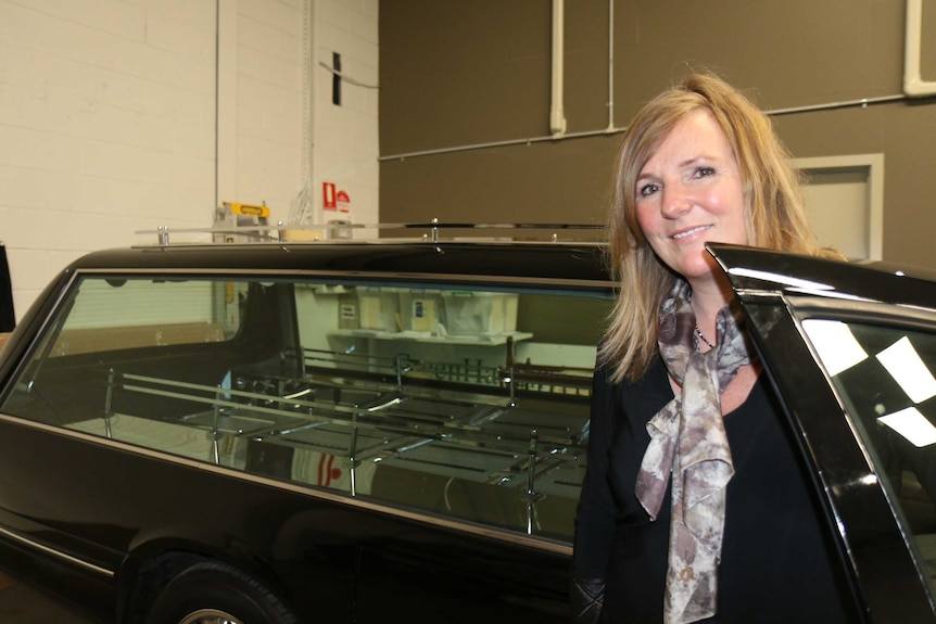Carly Dalton stands smiling next to a large black hearse.
