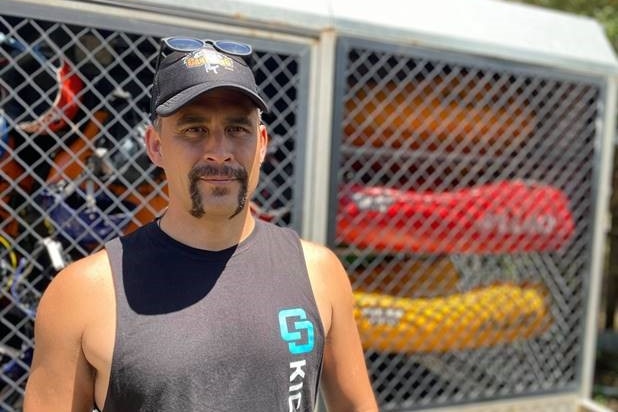 A middle-aged muscular man in cut-off T-shirt stands in front of shed on a sunny day.