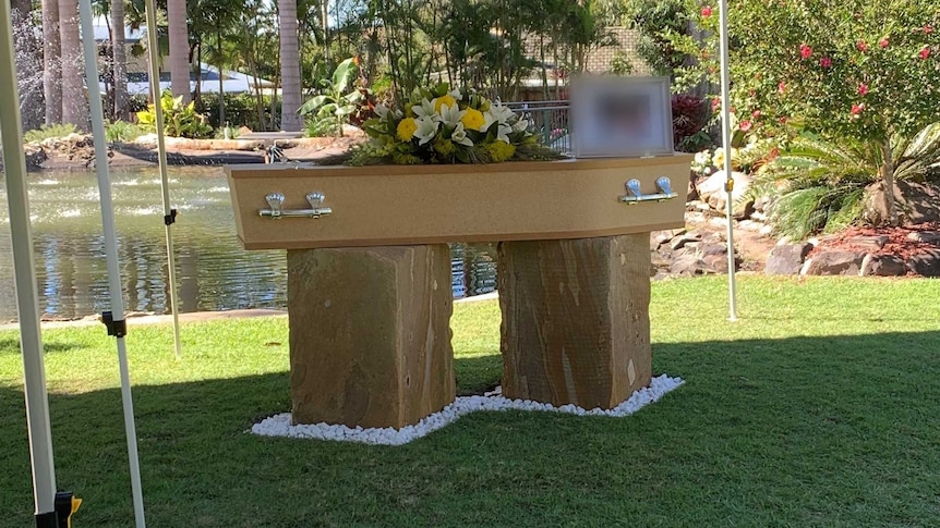 A coffin with flowers on top sits under a marquee outside with a pond in the background