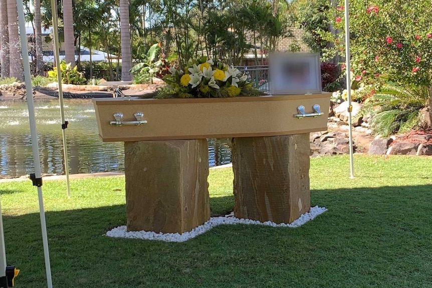 A coffin with flowers on top sits under a marquee outside with a pond in the background