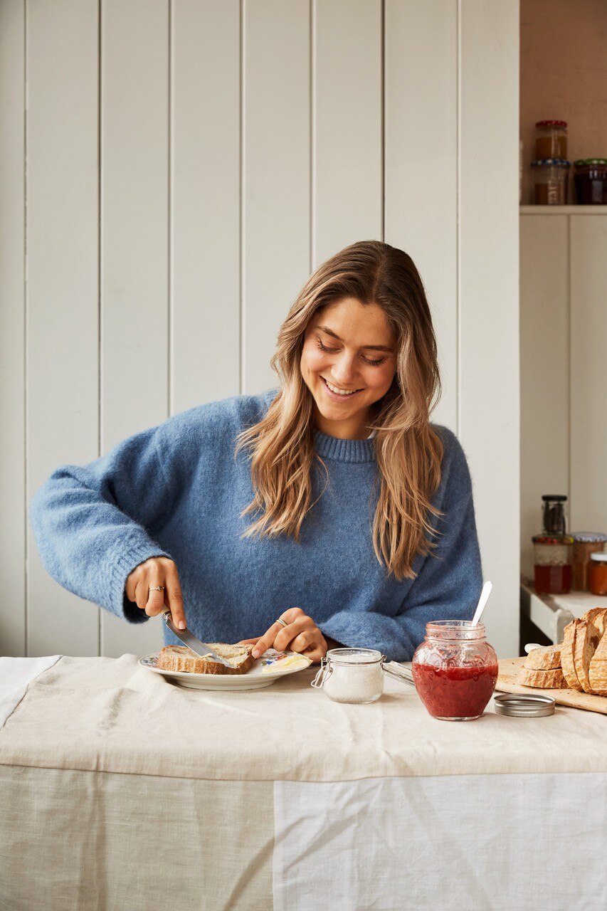 Claire Dinhut, smiling, spreads butter on a piece of bread. A jar of butter and a jar of red jam sit on the table.