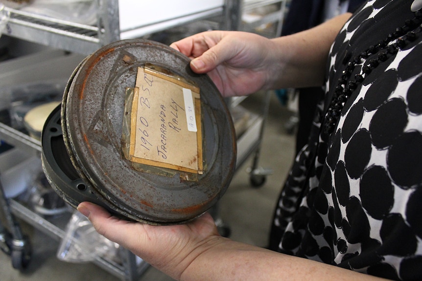 A woman's hands holding a film reel in a rusted metal tin, labelled "1960 Jacaranda Rally".