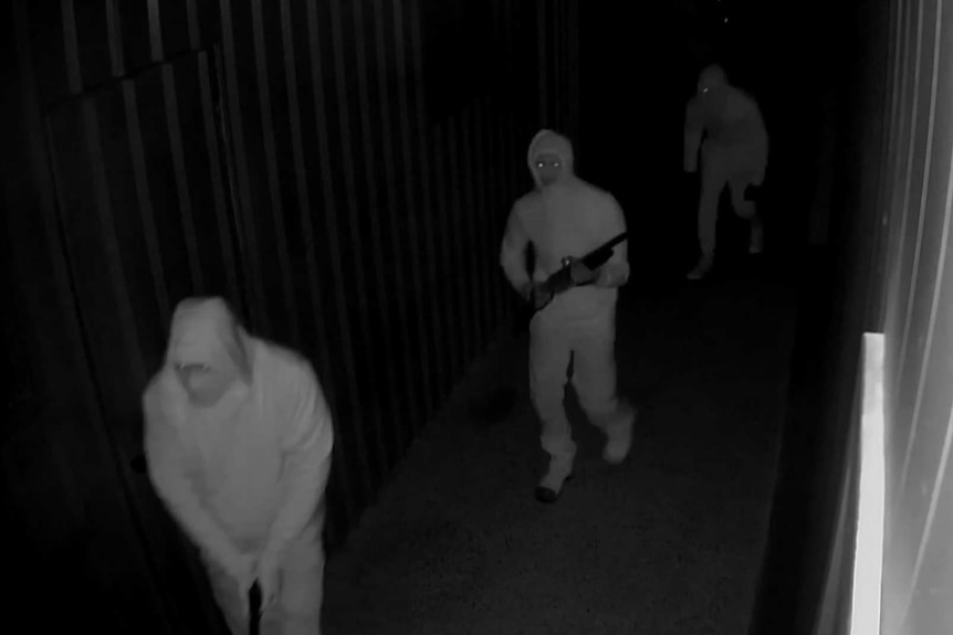 Three hooded men appear in a black and white screengrab. Two of them are carrying guns.